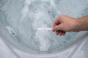 A hand holds a water test strip above the churning water of a backyard hot tub jet