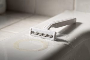 A used disposable razor with hairs caught in the blades lies in soapy water on a bathtub edge