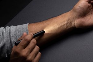 A penlight held flat against a forearm rakes light across the skin, revealing fine hairs and texture