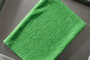 A green Korean exfoliating mitt of crinkled viscose lies on a steel tray, side light catching every ridge